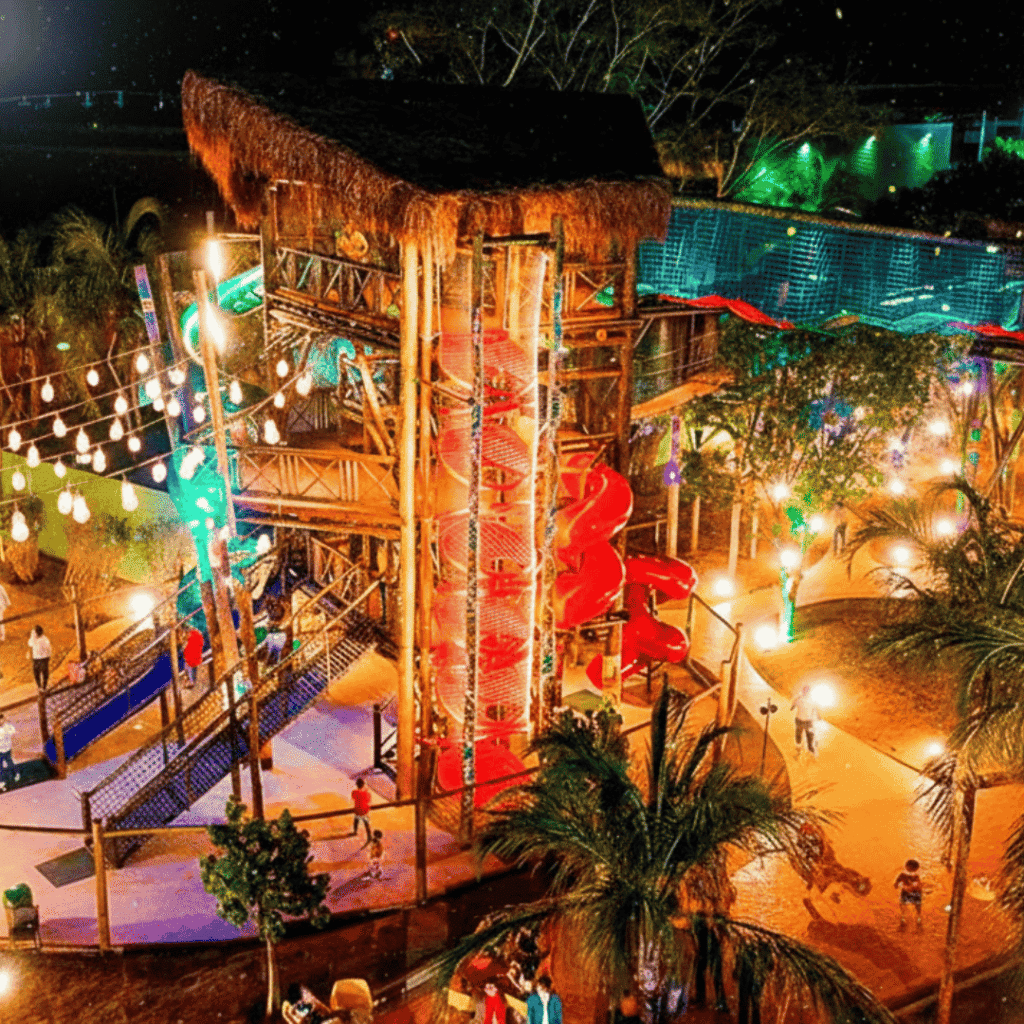 Vista noturna da Vila Guarani em Olímpia, com iluminação vibrante, brinquedos interativos e área de lazer familiar cercada por palmeiras e clima acolhedor.