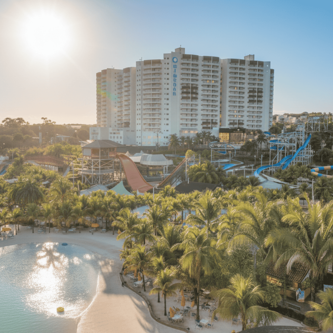 Vista completa do Wyndham Olímpia Royal Hotels ao lado do Thermas dos Laranjais, com a praia artificial, as atrações aquáticas e as fileiras de palmeiras iluminadas pelo pôr do sol.